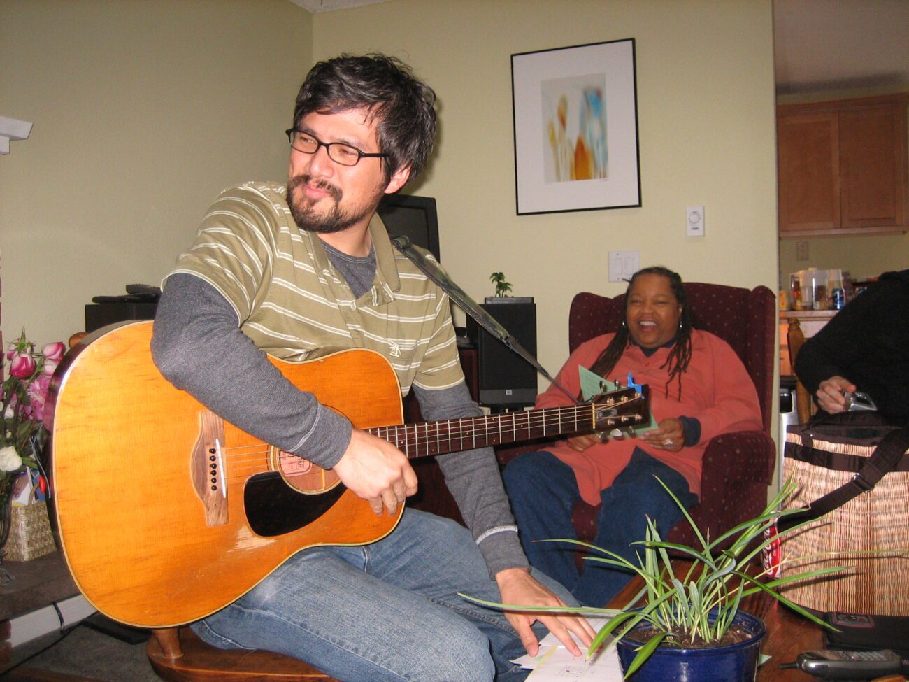 Michael Mathewson is seated on a chair at a gathering in someone's home playing a guitar strapped around his neck