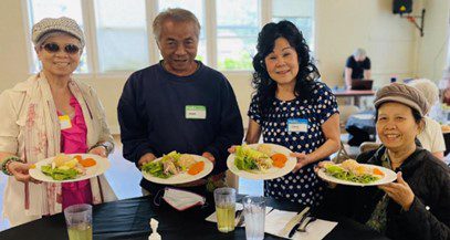 Four older adults of color hold up plates full of bright, nutritious foods at their table during a community meal at South Park Senior Center.
