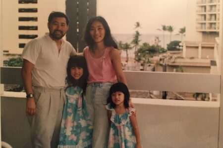 Mari Sugiyama with her father, mother, and sister on a family trip to Hawaii as a child, on a deck at a hotel with palm trees, sand, and ocean seen in the background