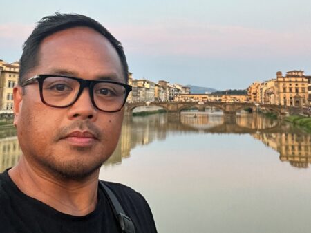 Selfie image of Edwin Obras with a bridge over a river and and buildings in the distance in Florence, Italy.