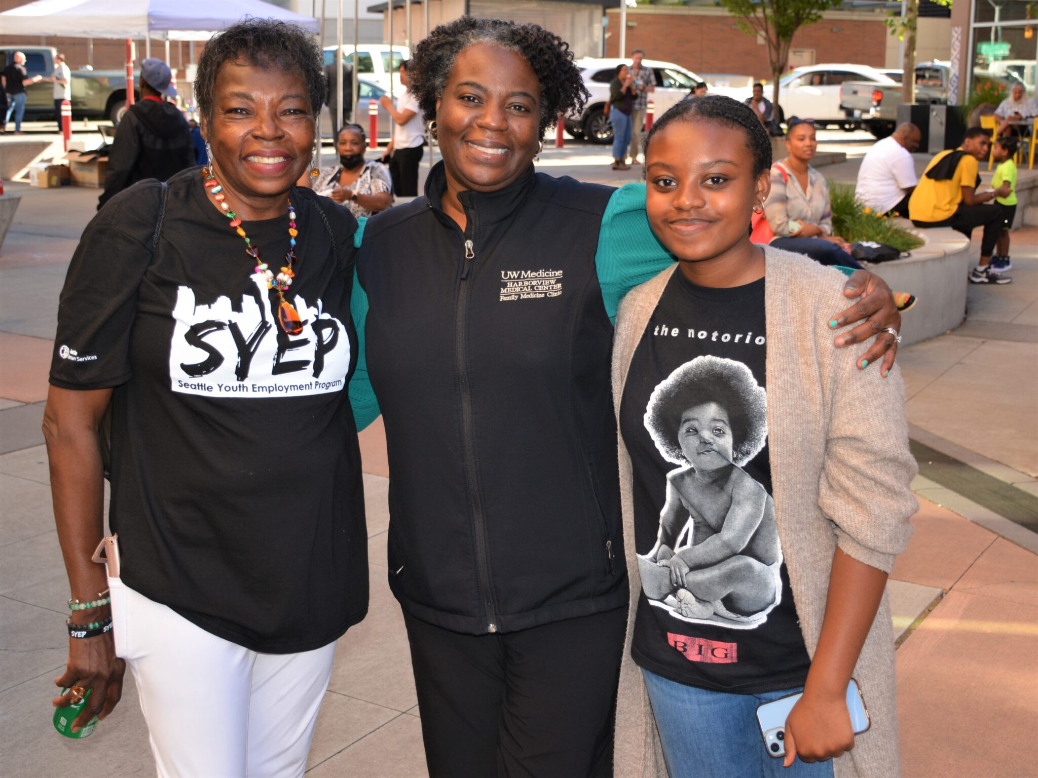 Three Black women, a grandmother, mother, and daughter, smile for the camera. the grandmother is wearing a T-shirt with a SYEP logo on it.