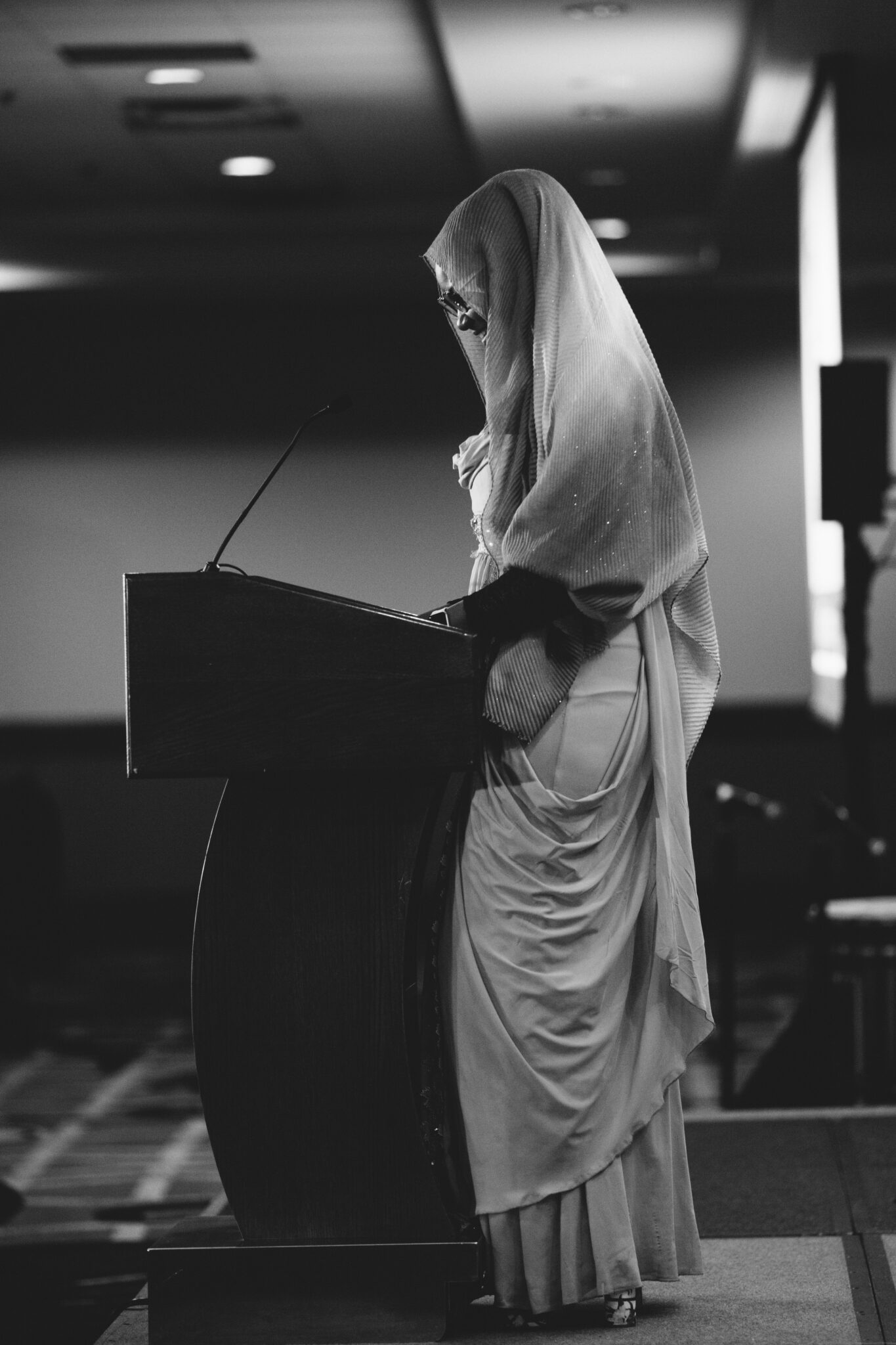 A young woman in traditional Muslim clothing stands smiling at a podium reading a speech.