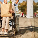 grocery shopper who uses wheelchair holds a large bag of groceries on her lap while companion pushes chair