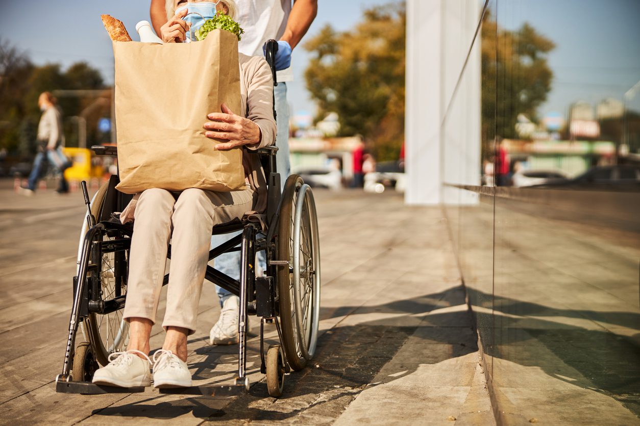 grocery shopper who uses wheelchair holds a large bag of groceries on her lap while companion pushes chair