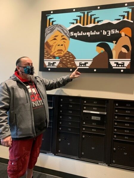 Man standing in a permanent housing building mailroom and pointing at native art hung on the wall above