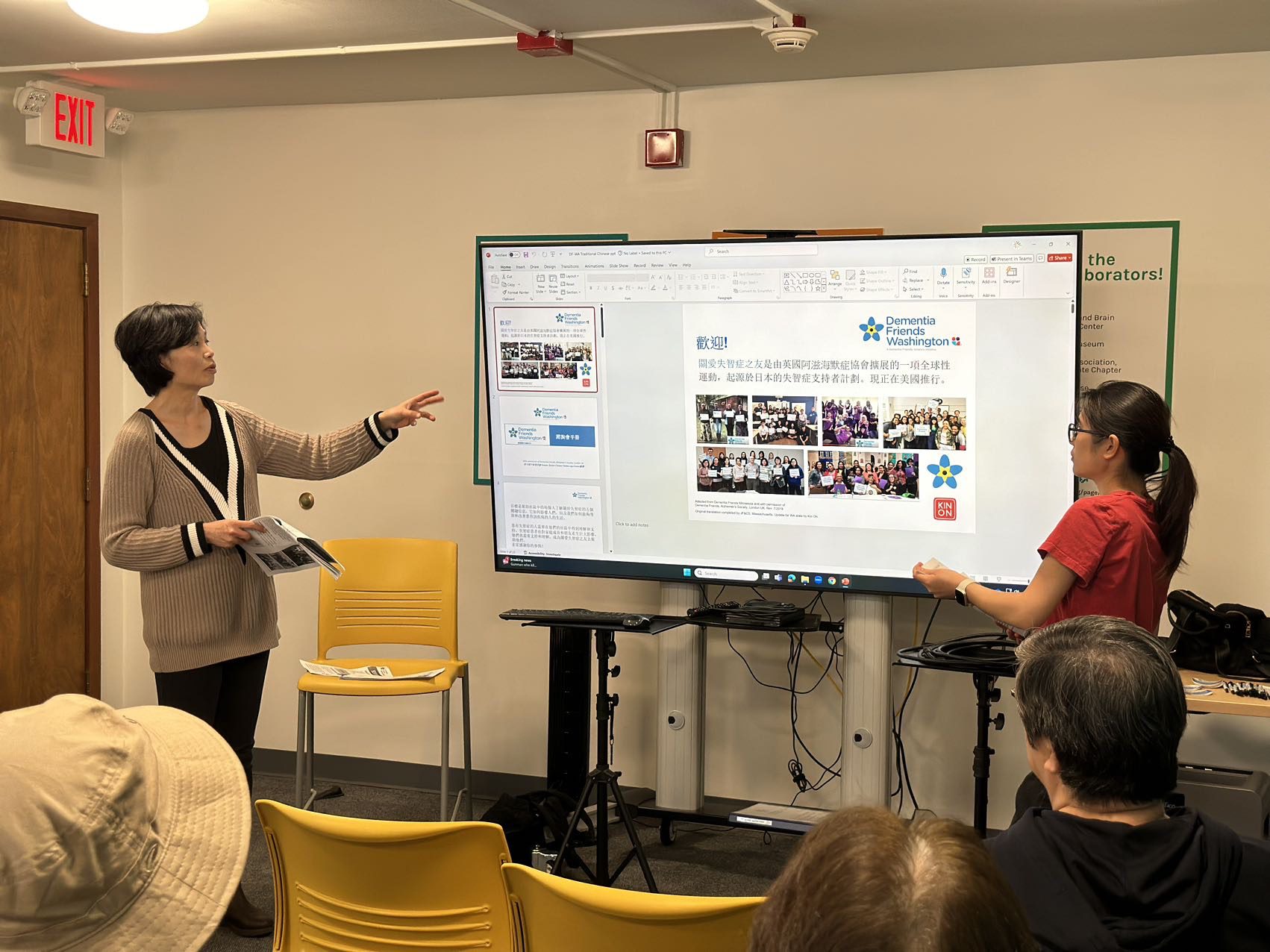 Two women in front of a large screen leading a presentation about Dementia Friends Washington with slides presented in Chinese.