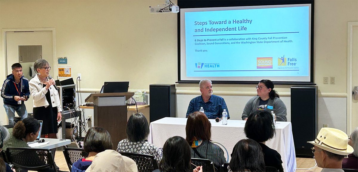Representatives of Sound Generations' Minor Home Repairs sit at a table during a PowerPoint presentation as a moderator speaks on a microphone.