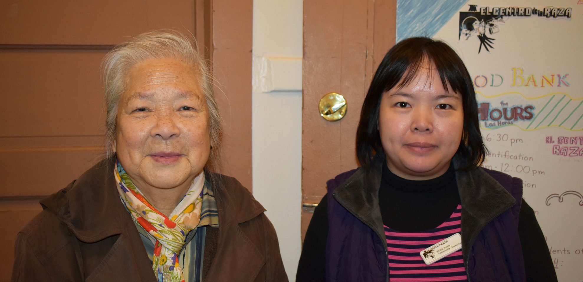 An older woman stands beside Katie Yuen in front of a Food Bank whiteboard.