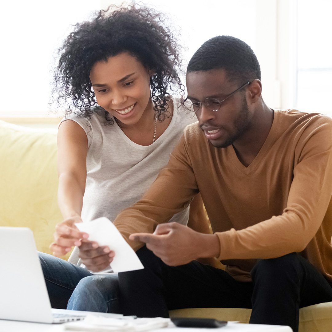 A smiling couple looks at a utility bill in front of a laptop computer.